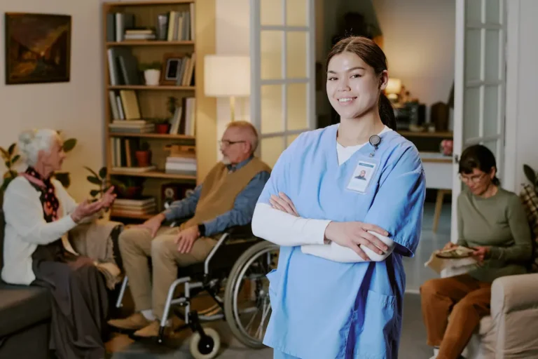 Young adult Hispanic woman nurse standing with arms crossed smiling at camera, senior Caucasian man in wheelchair and senior Caucasian women sitting and talking in background in care facility